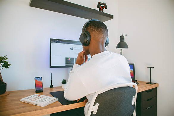 Homem negro sentado de costas para a câmera olhando para a tela do computador. Ele está usando uma camiseta branca de manga longa. A ideia da imagem é mostrar como os hábitos influenciam na rotina.